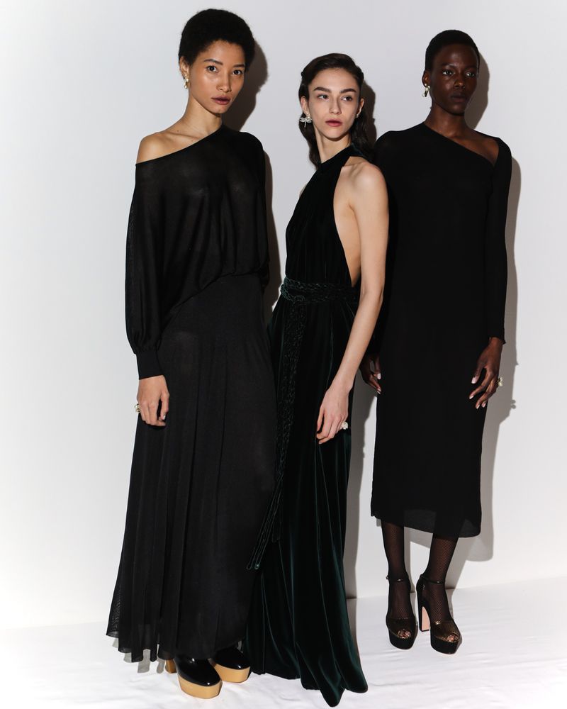 Three models stand in a minimalist studio wearing black dresses, showcasing varied designs and elegant silhouettes in a fashion editorial.