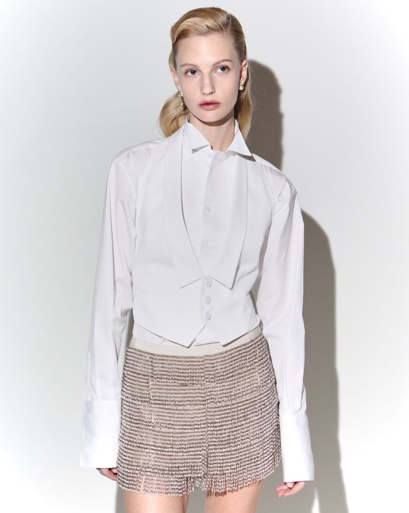 Woman poses in a white shirt and embellished fringe mini skirt against a white backdrop in a fashion studio shot with bright, even lighting.