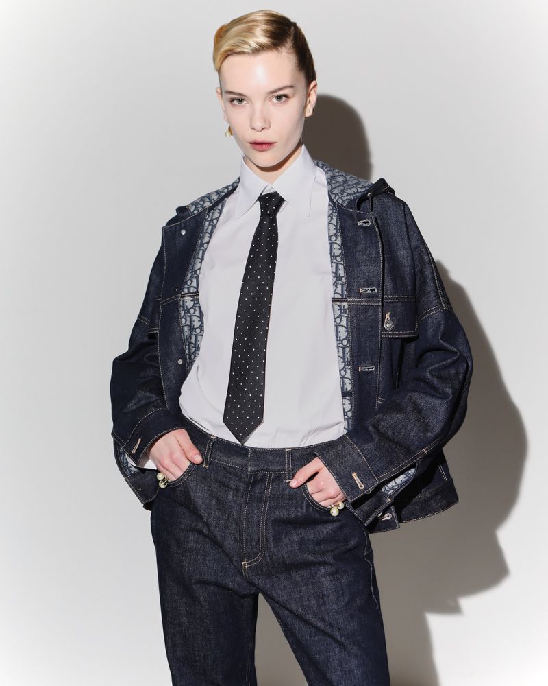 A blonde woman poses in a dark blue denim jacket and pants, wearing a white collared shirt and a polka dot tie against a white backdrop.