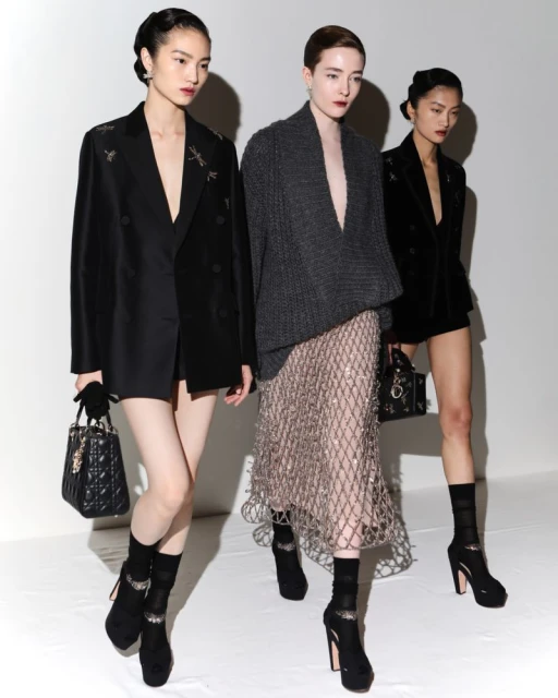 Three fashion models pose in studio wearing blazers, skirt, knit shawl, and platform heels, each carrying a small black purse.