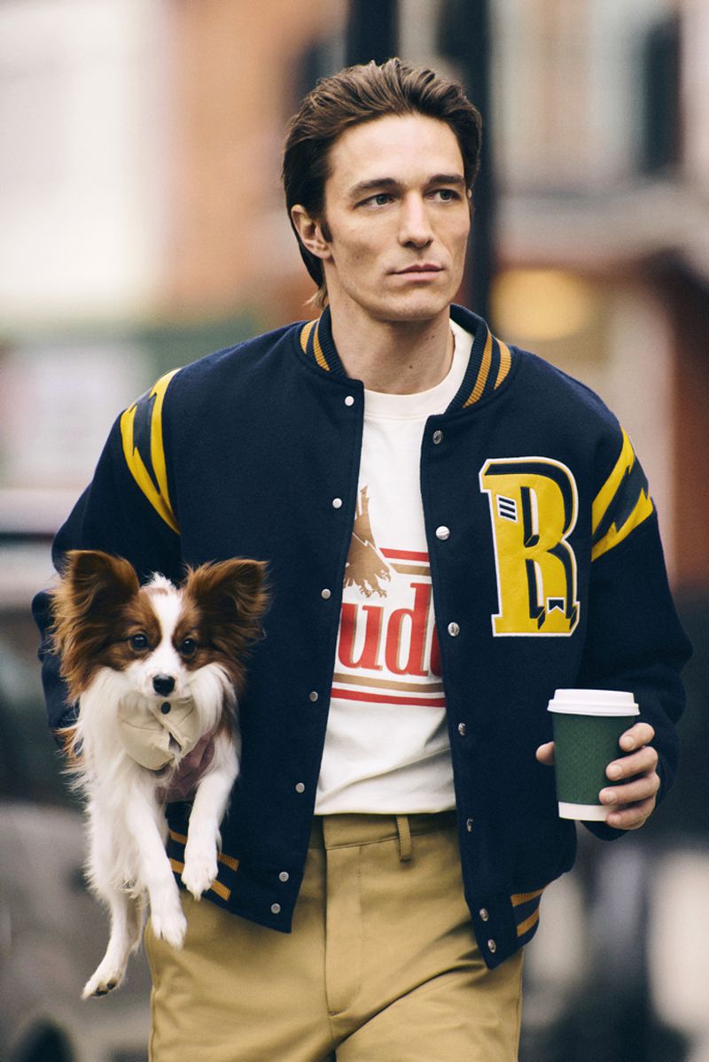 An adult male in a letterman jacket holds a small dog, walking outdoors while holding a paper coffee cup, showcasing fashion and lifestyle.