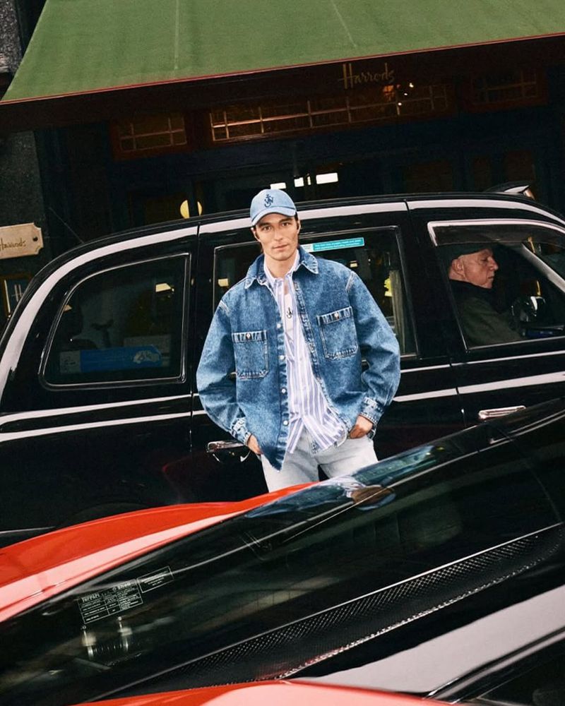 Man poses near black taxi and luxury car outside Harrods department store wearing denim jacket, striped shirt, and baseball cap.