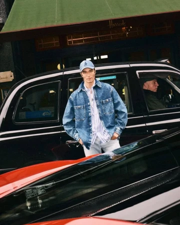 Man poses near black taxi and luxury car outside Harrods department store wearing denim jacket, striped shirt, and baseball cap.