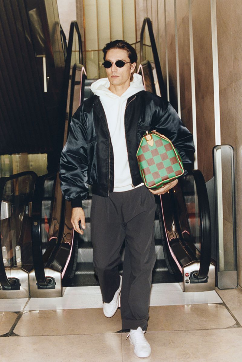 A stylish adult male descends an escalator wearing a jacket, white hoodie, sunglasses and trousers, and carrying a patterned duffel bag.