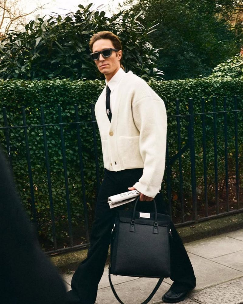 Man in sunglasses walks on a sidewalk holding a black tote bag and newspaper with a hedge and fence in the background.