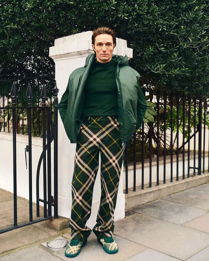 Adult male poses outdoors near fence wearing green turtleneck, puffer vest, plaid pants, and sneakers, with green foliage background.