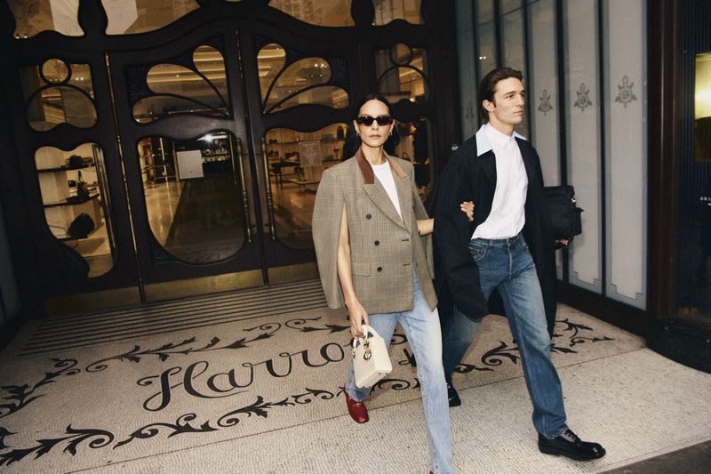 A stylish woman and man walking in front of Harrods, the woman in a blazer and the man with a jacket draped over his shoulder.