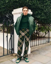 Man wearing a green puffer vest and plaid trousers standing outside near a black metal fence and white pillars