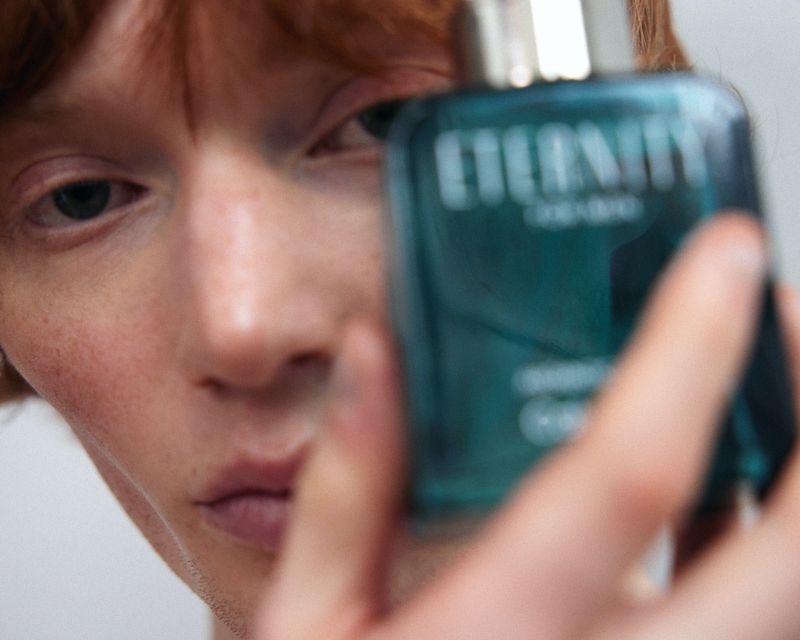 Close-up studio shot of a red-haired male adult holding a green glass Eternity cologne bottle against a plain white backdrop.