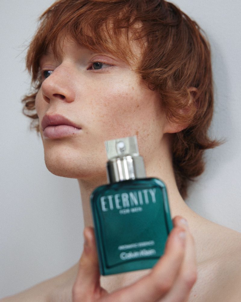 Close-up studio shot of a young adult male holding a green 'Eternity for Men' cologne bottle, promoting the fragrance with a clean, modern aesthetic.