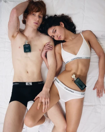 Two models, a man and a woman, laying on a bed with Calvin Klein underwear and Eternity perfume bottles, showcase a intimate studio shot.