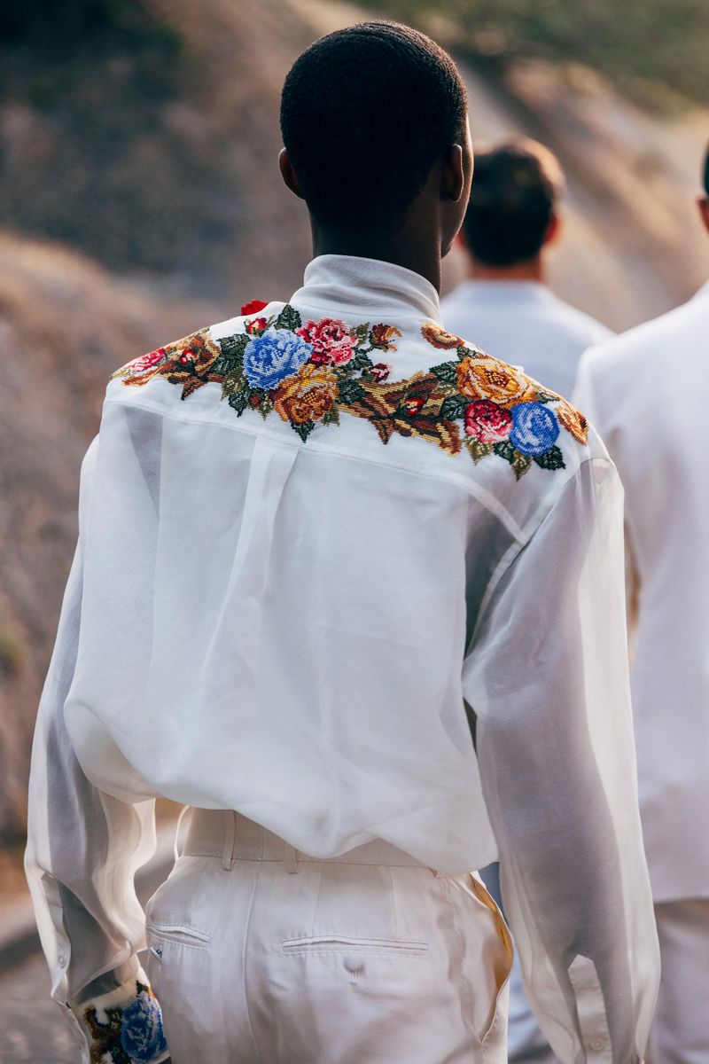 Person in a white shirt with floral embroidery on the back walks with other people in matching outfits outdoors during daytime.