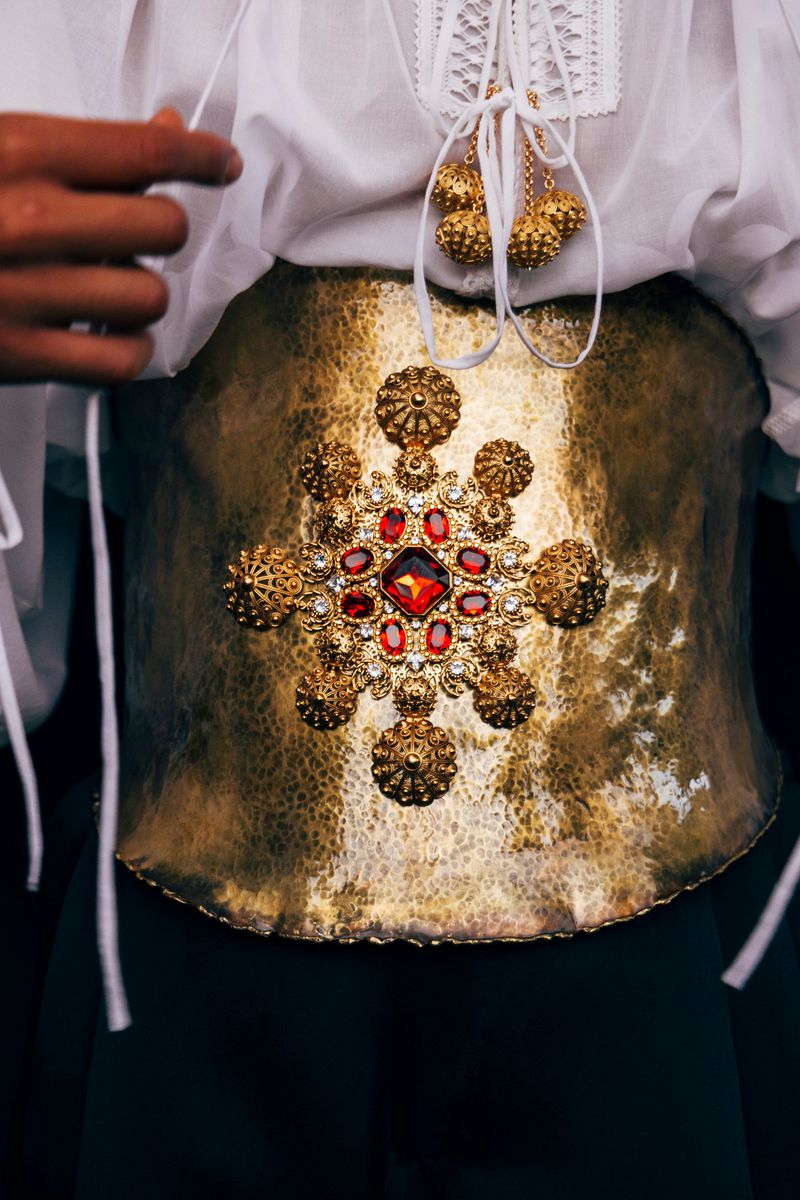 Close-up of a person wearing a decorative, hammered-gold girdle with red gemstones, with a white lace shirt and dark skirt.