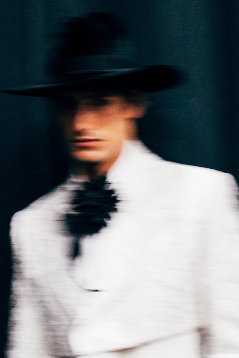 Artistic portrait of an adult male wearing a wide-brim hat and white jacket against a dark background, blurred motion effect.
