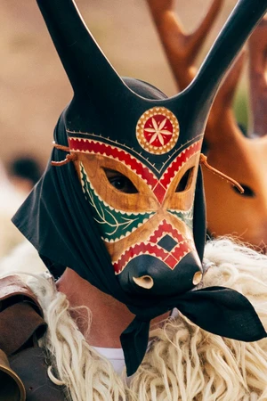 Person wearing an ornate carved wooden horned animal mask with red and green patterns and thick white shaggy fleece coat for Dolce & Gabbana