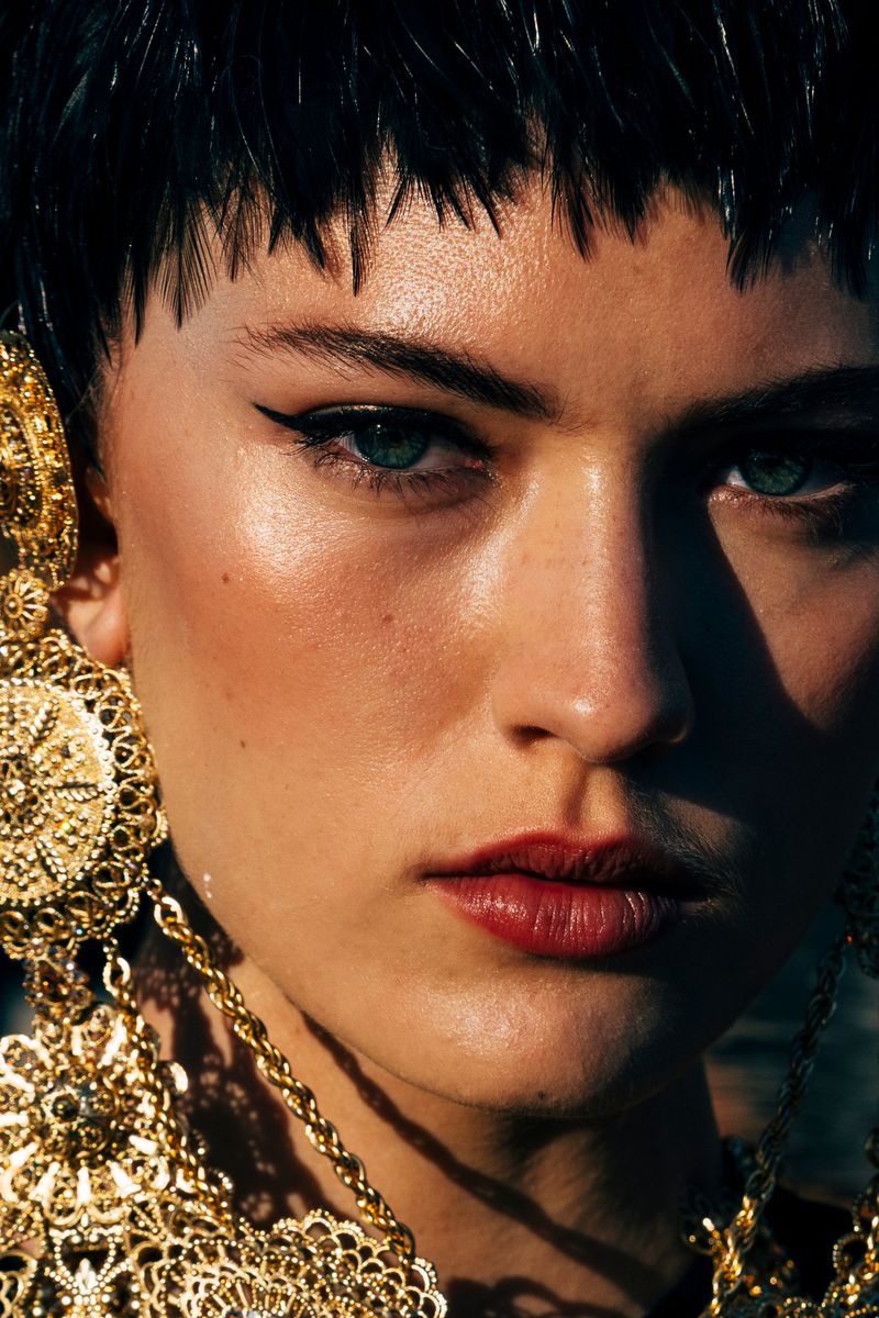 Close-up portrait of a woman with short dark hair wearing ornate gold filigree earrings for Dolce & Gabbana Alta Moda Fall 2024.