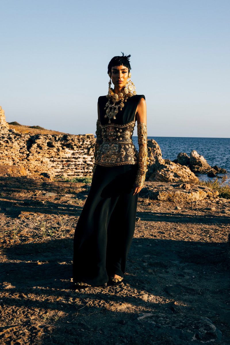 Woman in ornate black and gold gown stands on rocky shore with the ocean in the background under clear sky. Fashion, editorial.