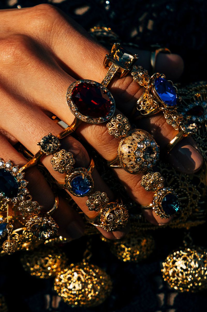 Hand adorned with multiple gold rings featuring red and blue gemstones, set against a dark, glittering textile backdrop in warm light.