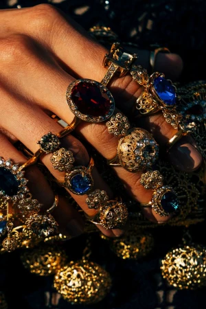Hand adorned with multiple gold rings featuring red and blue gemstones, set against a dark, glittering textile backdrop in warm light.