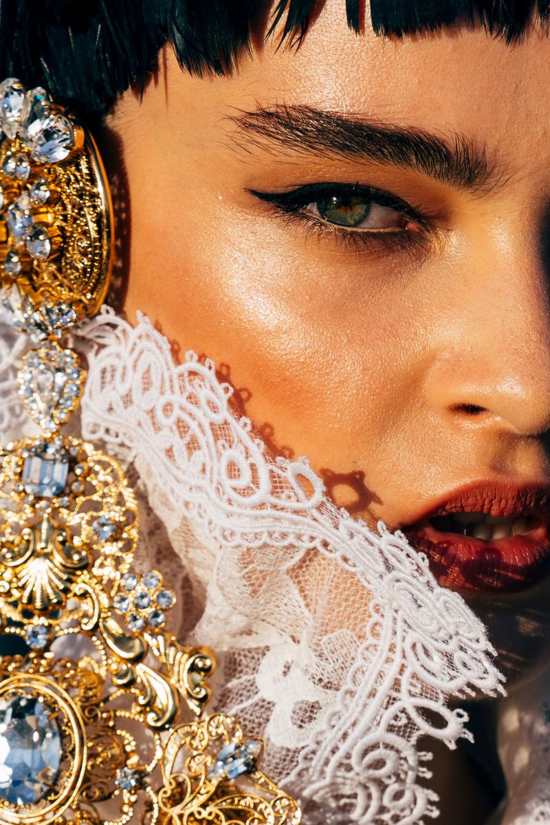 Close-up portrait of a model with dark hair wearing heavy gold and jeweled jewelry, a high lace collar, and bold makeup.