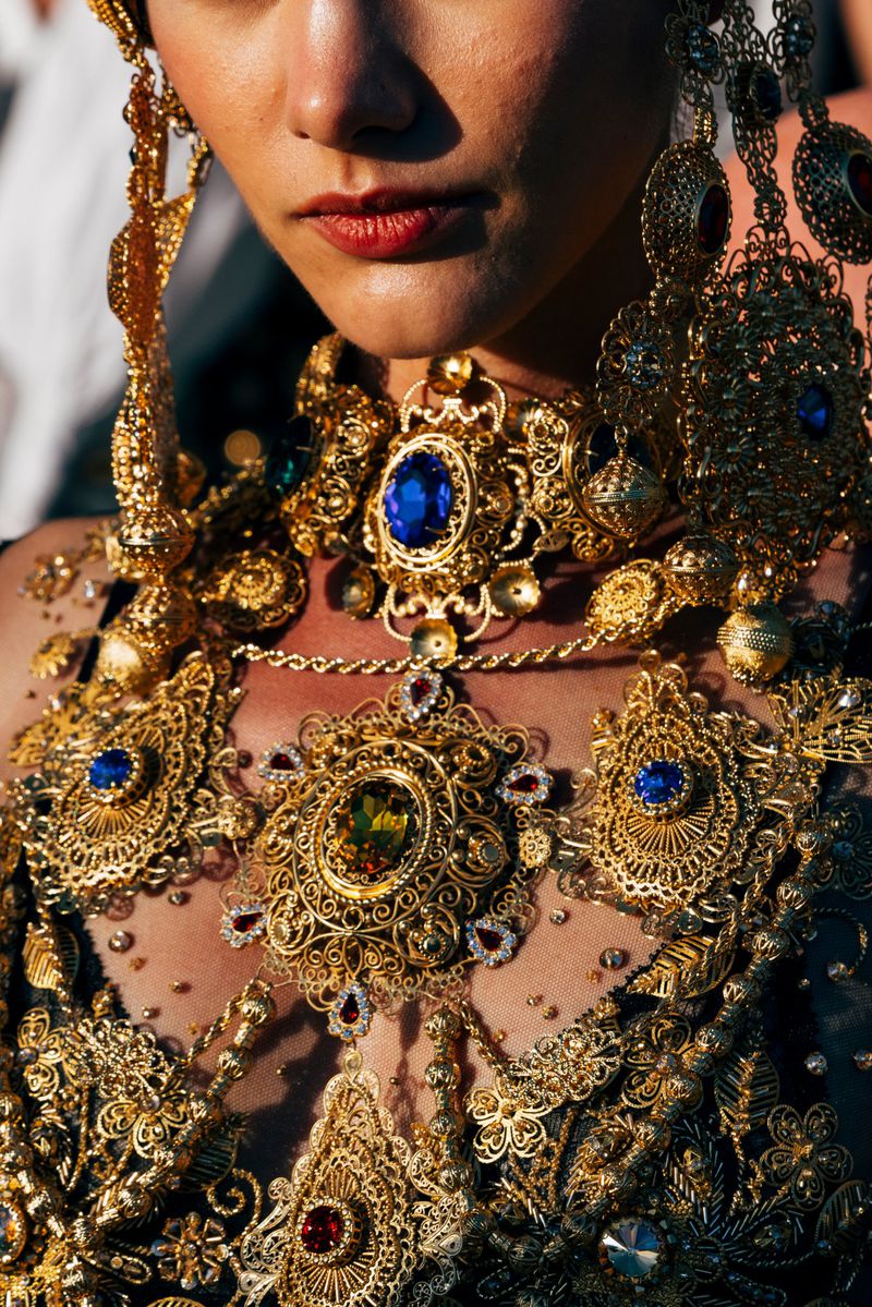 Close-up of a woman adorned with intricate gold jewelry, including a large necklace with a blue gemstone, and elaborate headpiece.