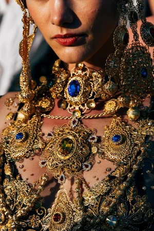 Close-up of a model wearing ornate gold filigree jewelry with blue and red gemstone accents for Dolce and Gabbana Alta Moda.