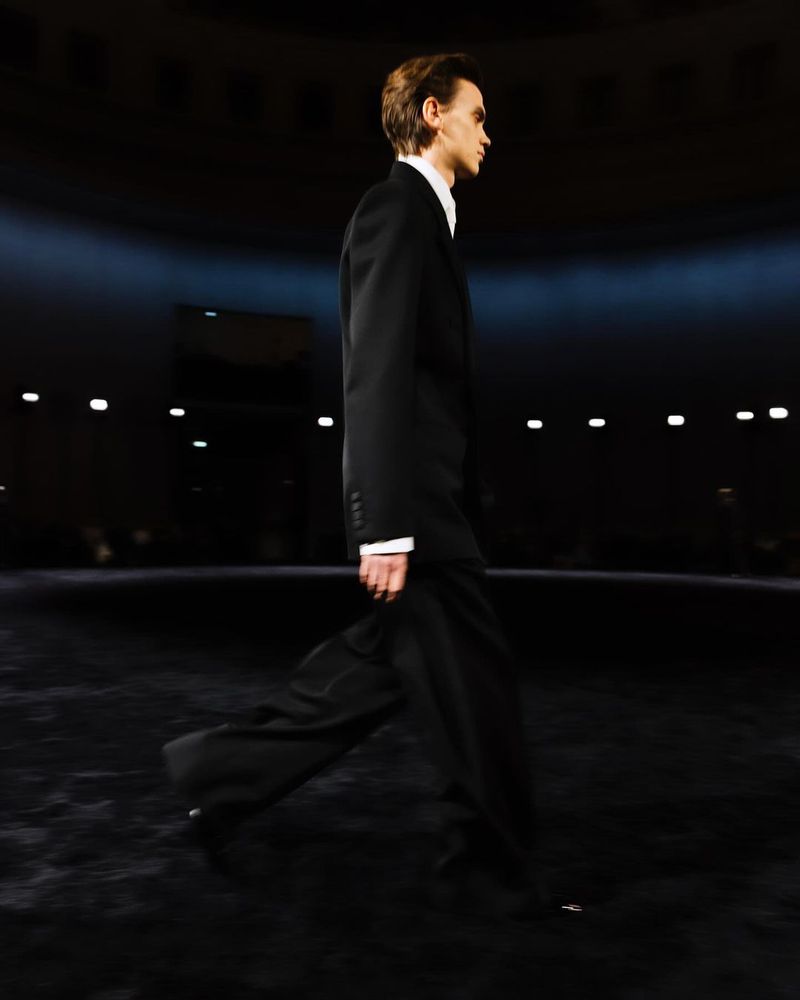Male fashion model walks the runway in a dark indoor venue wearing a black suit and white dress shirt, side view, low lighting.