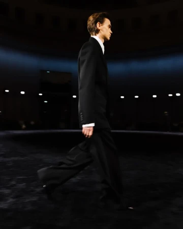Male fashion model walks the runway in a dark indoor venue wearing a black suit and white dress shirt, side view, low lighting.