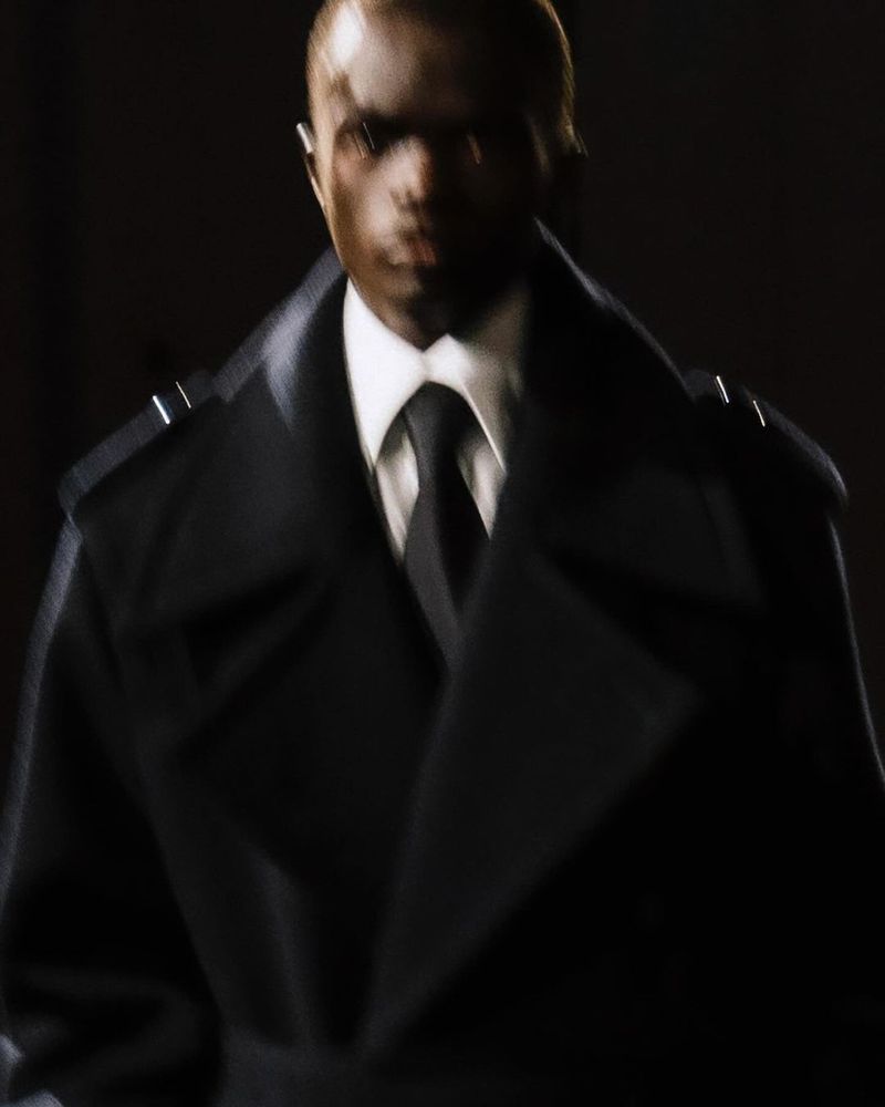 Close up of an adult Black male wearing a dark overcoat, collared shirt, and tie, looking forward in a dimly lit indoor setting.