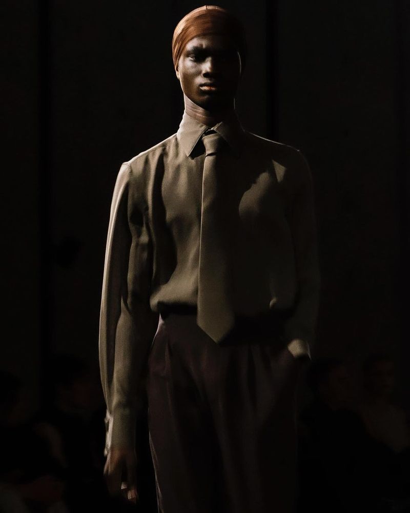 Portrait of an androgynous adult wearing a neutral-colored shirt, tie, and slacks against a dark background with stark lighting.