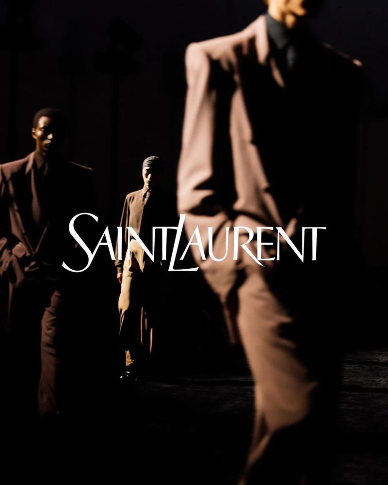 Three fashion models wearing suits walking in the dark with the Saint Laurent logo visible in the foreground.