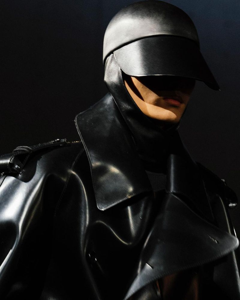 Person in black shiny coat and headpiece with visor, creating a futuristic and mysterious fashion statement against a dark backdrop.