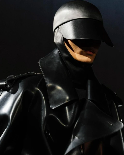 Person in black shiny coat and headpiece with visor, creating a futuristic and mysterious fashion statement against a dark backdrop.