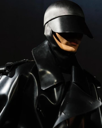 Person in black shiny coat and headpiece with visor, creating a futuristic and mysterious fashion statement against a dark backdrop.