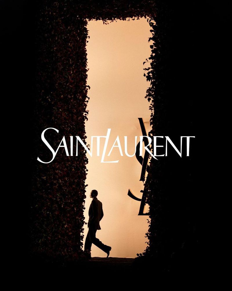 Person walks through a plant-lined frame showcasing the Saint Laurent brand against an orange backdrop, fashion-forward, silhouette.