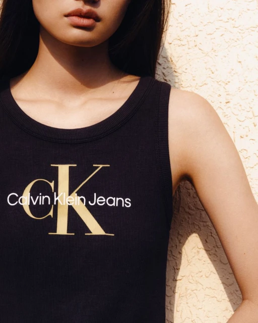 Close-up of a light-skinned woman wearing a black Calvin Klein Jeans tank top with gold logo, softly lit against a textured cream background.