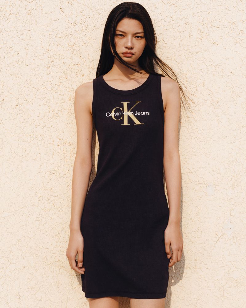 Woman in a black tank dress with Calvin Klein Jeans logo stands against a textured wall in a medium shot, fashion forward and trendy.