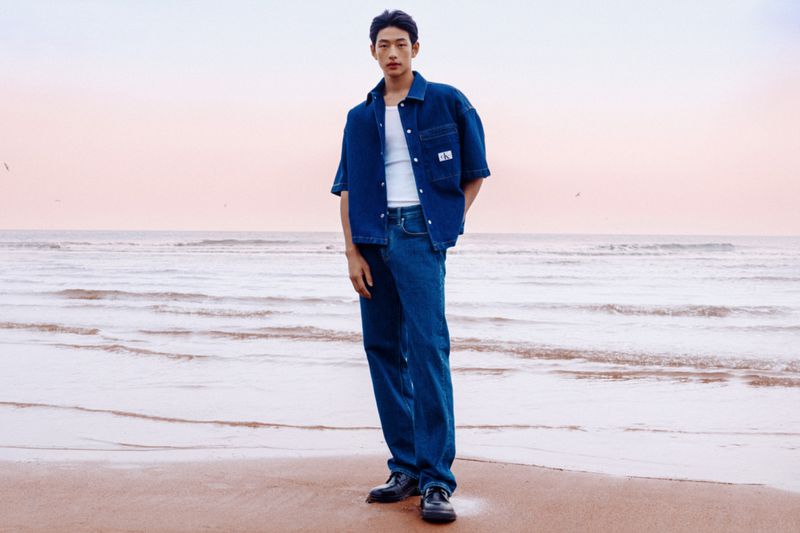 East Asian male model wearing a denim shirt and jeans standing on a sandy beach at sunset for Calvin Klein.