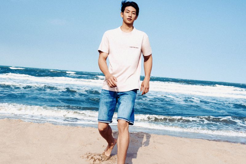 Adult male stands barefoot on a beach in natural light, posing in jeans shorts and a t-shirt with the ocean and sky in the background.
