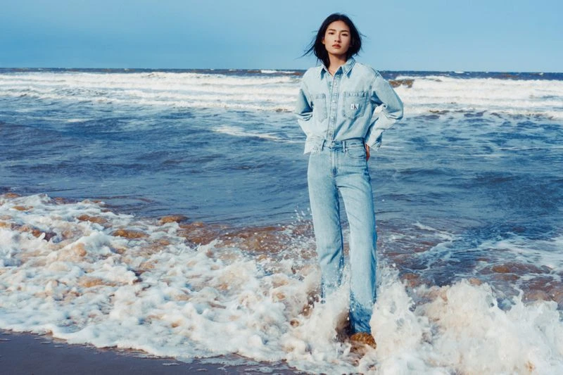 Woman in denim outfit stands on beach with ocean waves. Light blue denim shirt and jeans, dark hair, outdoor shot, ocean background.