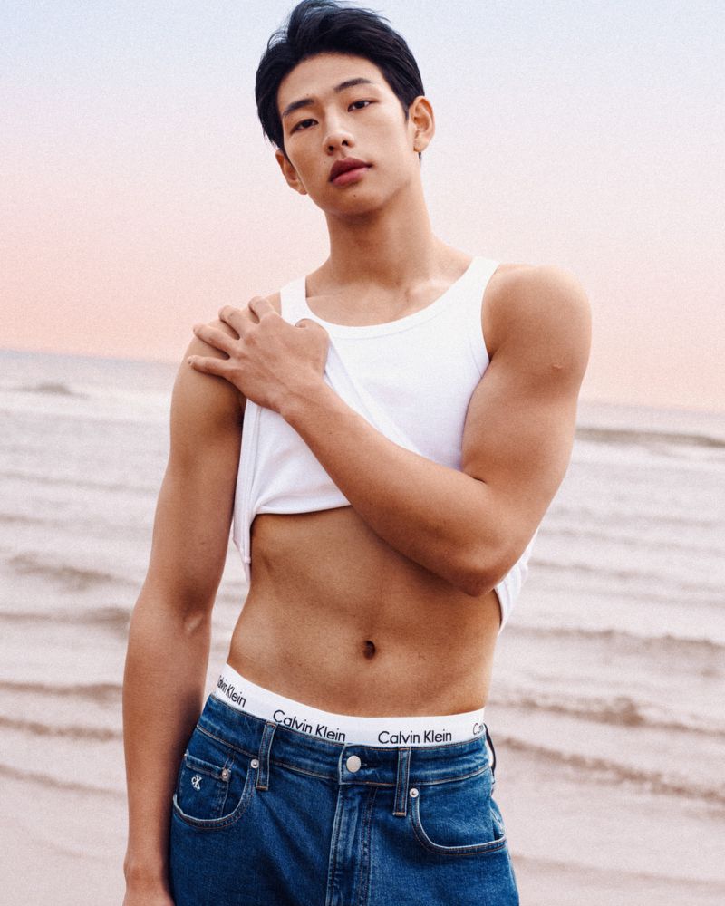 Attractive Asian man in a white tank top and blue jeans poses against a beach background with soft, natural lighting and sky.