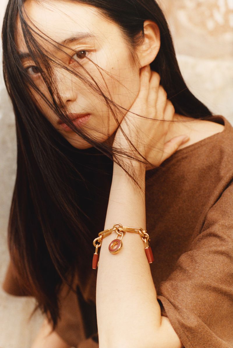 Portrait of a young Asian woman in a brown top, gold charm bracelet on her wrist, and dark hair partially covering her face.