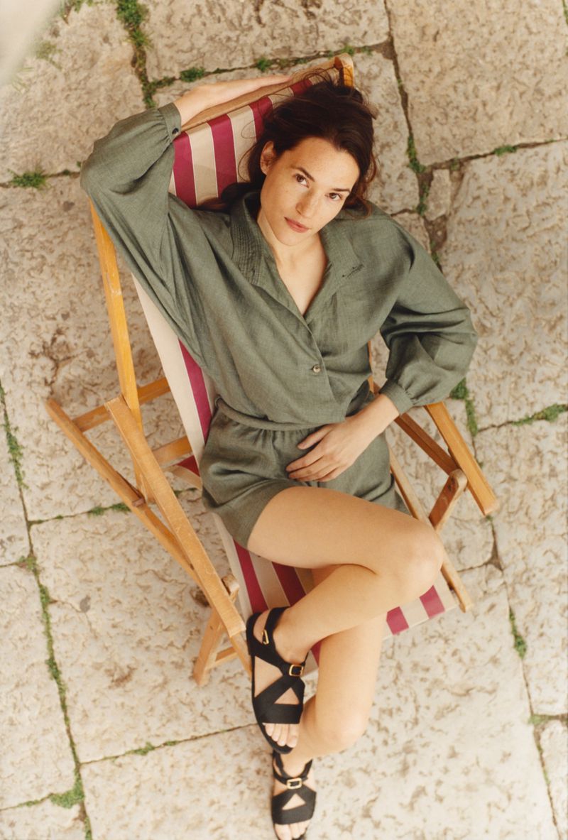 Brunette woman in an olive green shorts outfit reclines in a striped beach chair on stone pavers, shot from an overhead high angle.
