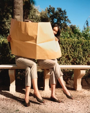 Two Asian women sit on a park bench, holding up a large sheet of brown paper that obscures their faces, creating a playful, candid outdoor scene.