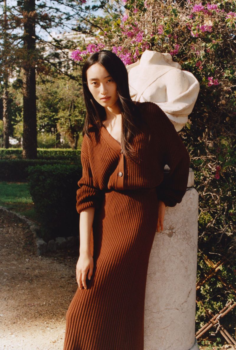 A stylish woman poses in a brown knit dress and cardigan in a sunlit garden with a classic marble bust and blooming pink flowers.