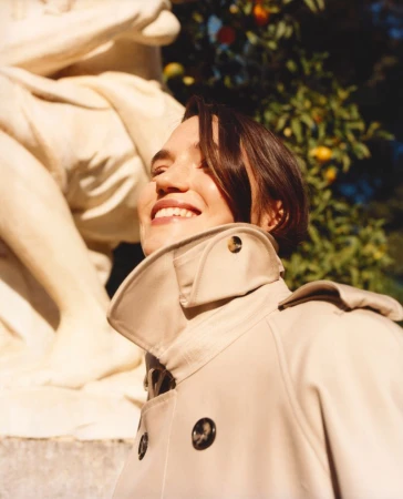 A smiling woman with short brown hair in a tan trench coat stands in front of a marble sculpture and a tree with orange fruit.