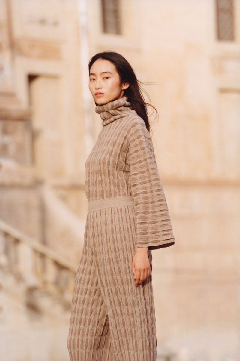 Portrait of a slender Asian woman in a neutral-tone knit outfit, posing outdoors against an out-of-focus architectural backdrop.