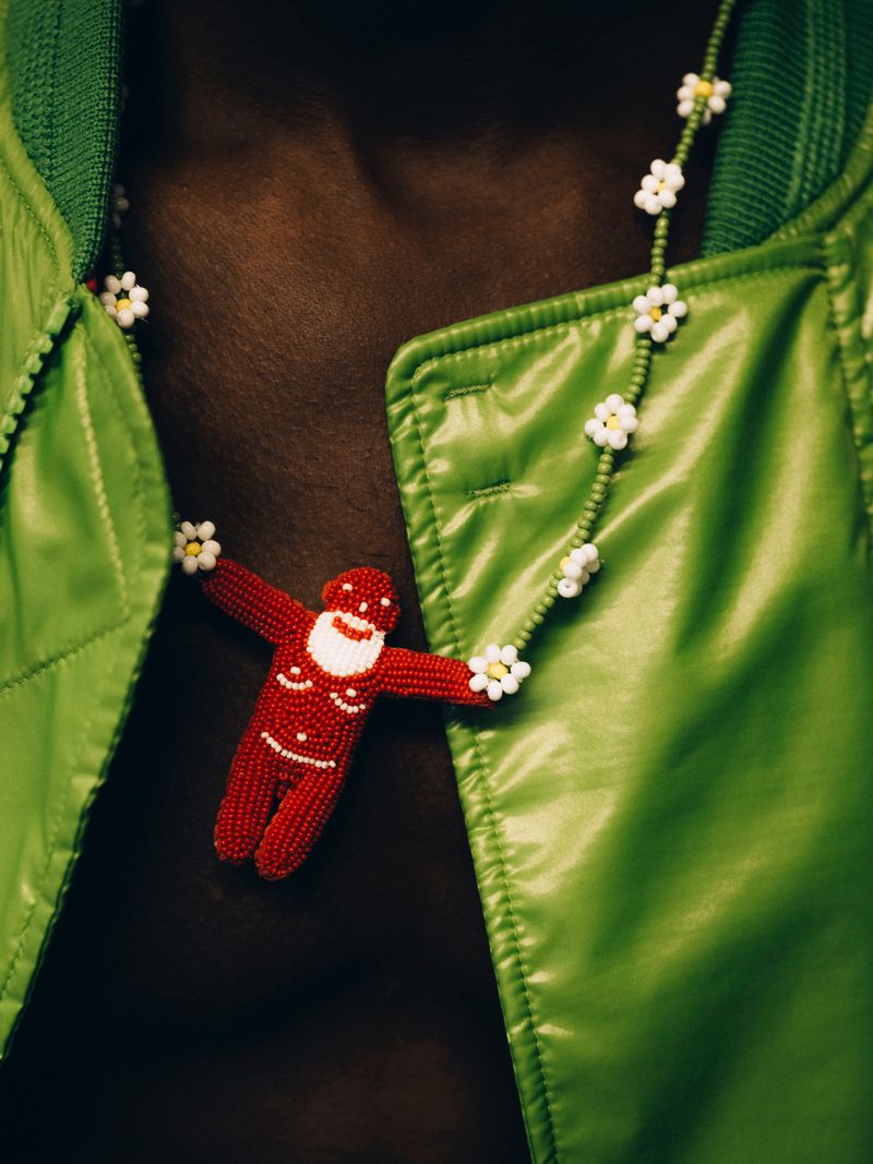 Close-up studio shot of a dark-skinned torso wearing a green jacket, a beaded daisy necklace, and a red beaded Santa Claus pendant.