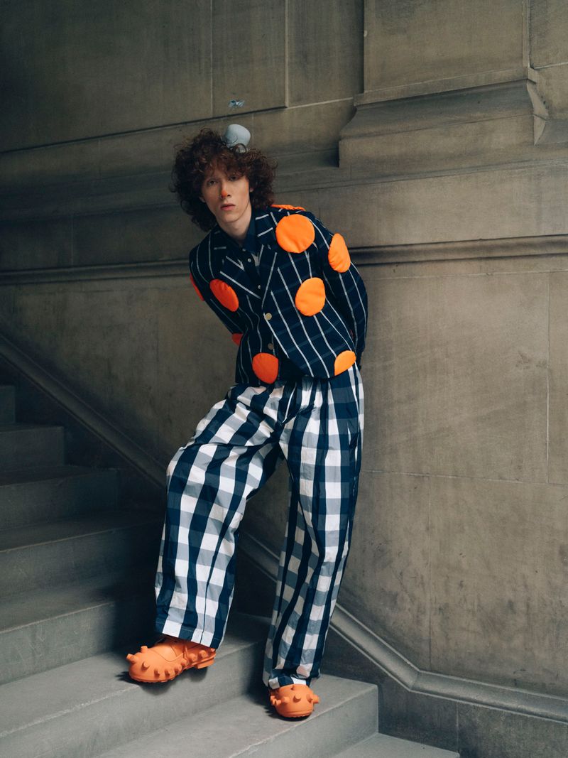 Adult fashion model poses on stairs wearing a striped jacket with orange dots, checkered pants, and orange spiky shoes.