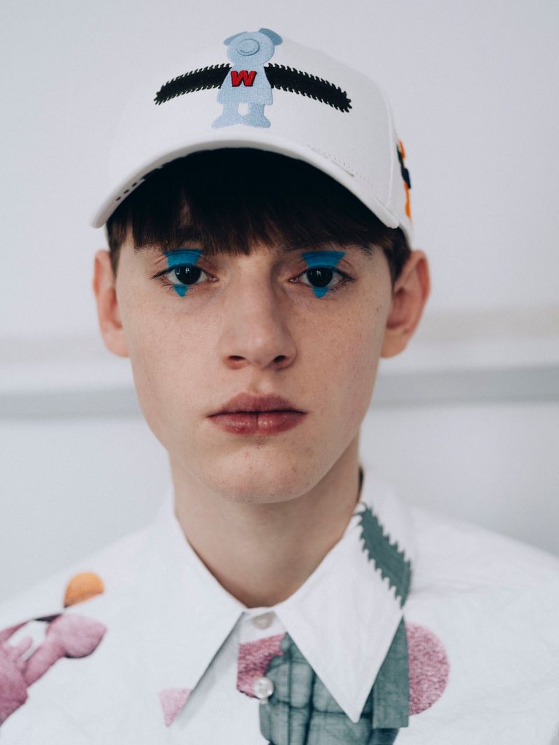 Close-up of a person with blue makeup under their eyes, wearing a white baseball cap with a cartoon bear, and a collared shirt.